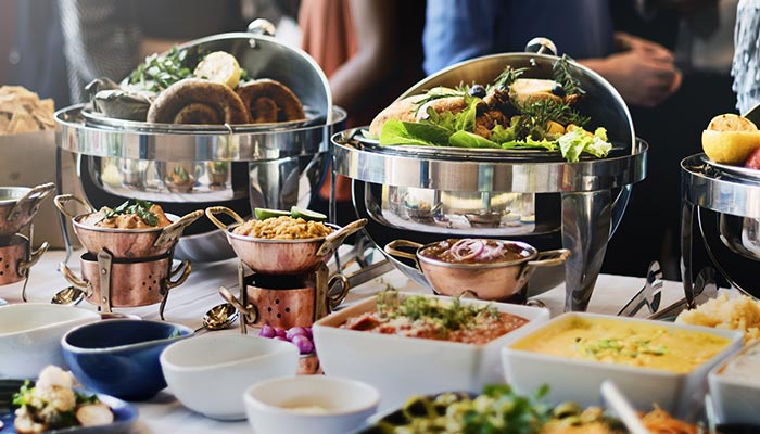 A restaurant buffet table with breakfast, lunch & dinner dishes in trays, people in the background.